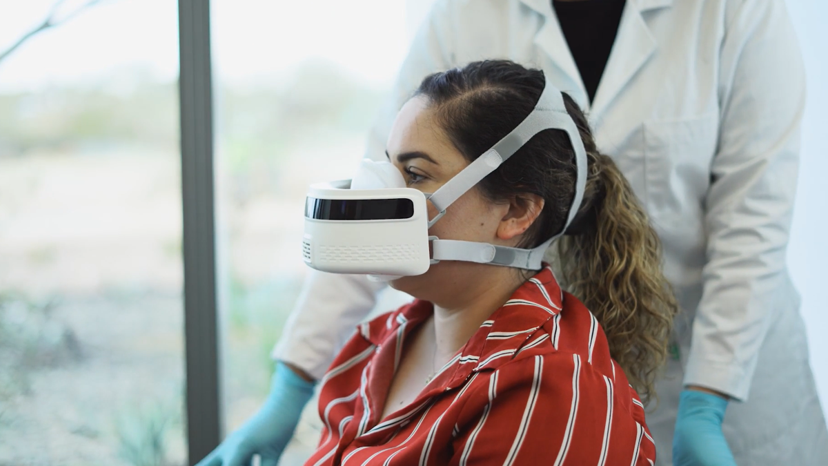 A woman wears the Breezing device on her face while a medical professional in a lab coat helps her.