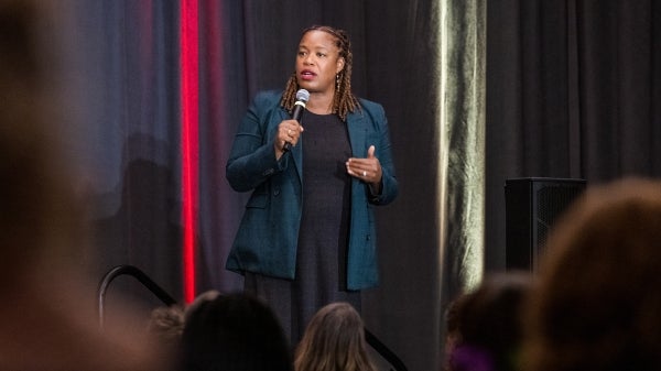 Black woman with medium length braided hair wearing a black dress and a teal blazer talks into a microphone in front of an audience