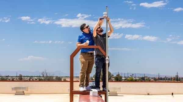 Two men install a temperature and humidity tower on a rooftop.