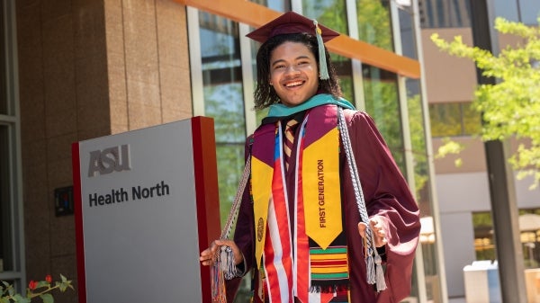 Dallas Salas smiles at the camera wearing his graduation cap and gown with all of his honor cords and stolls. He is sanding next to sign that says ASU Health North