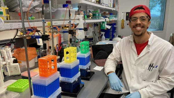 A man with a red snapback, a white lab coat, and blue gloves sits at a lab bench, smiling.